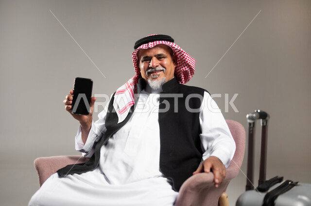 الاستعداد لقضاء وقت ممتع في الرحلات السياحية, بورتريه مقرب لرجل عربي خليجي سعودي مبتسم كبير بالسن يرتدي الثوب التقليدي والشماغ يجلس على الكرسي بجانب حقيبة الامتعة يحمل بيده الهاتف الجوال بشاشة فارغة, خلفية رمادية