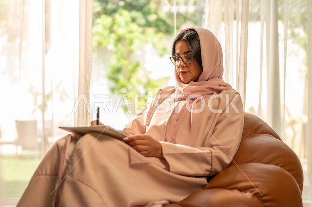 Acquiring artistic skills, passion and love for learning to draw, a young Saudi Gulf Arab woman sitting on a sofa in the living room and holding a notebook and pen in her hand, writing and preparing university assignments, the concept of education in the Kingdom of Saudi Arabia