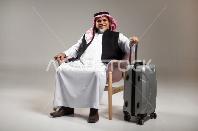 البحث والاستكشاف حول العالم, الاستعداد لقضاء وقت ممتع في الرحلات السياحية, بورتريه لرجل عربي خليجي سعودي مبتسم كبير بالسن يرتدي الثوب التقليدي والشماغ يجلس على الكرسي ويمسك بيده حقيبة الامتعة , خلفية رمادية