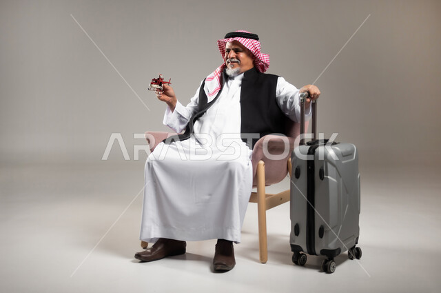 الاستعداد لقضاء وقت ممتع في الرحلات السياحية, مفهوم البحث والاستكشاف حول العالم, بورتريه لرجل عربي خليجي سعودي مبتسم كبير بالسن يرتدي الثوب التقليدي والشماغ يجلس بجانب حقيبة الامتعة ويحمل بيده مجسم طائرة حمراء, خلفية رمادية