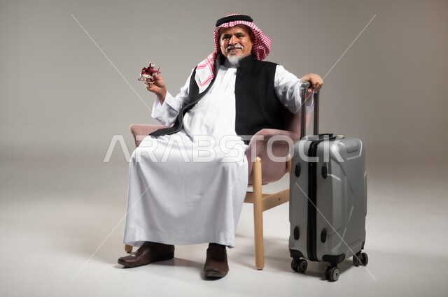 النظر الى الكاميرا بإيماءات الفرح و السرور, مفهوم البحث والاستكشاف حول العالم, بورتريه لرجل عربي خليجي سعودي مبتسم كبير بالسن يرتدي الثوب التقليدي والشماغ يجلس بجانب حقيبة الامتعة ويحمل بيده مجسم طائرة حمراء, خلفية رمادية