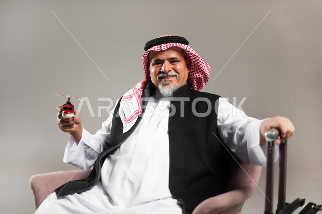 مفهوم البحث والاستكشاف حول العالم, بورتريه مقرب لرجل عربي خليجي سعودي مبتسم كبير بالسن يرتدي الثوب التقليدي والشماغ يجلس بجانب حقيبة الامتعة ويحمل بيده مجسم طائرة حمراء, النظر الى الكاميرا بإيماءات الفرح و السرور, خلفية رمادية