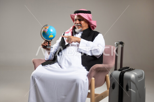 الاشارة إلى مكان معين على الخريطة والنظر إلى أحد القارات, مفهوم البحث والاستكشاف حول العالم, بورتريه مقرب لرجل عربي خليجي سعودي مبتسم كبير بالسن يرتدي الثوب التقليدي والشماغ يحمل بيده مجسم الكرة الارضية, الجلوس بجانب حقيبة الامتعة, خلفية رمادية