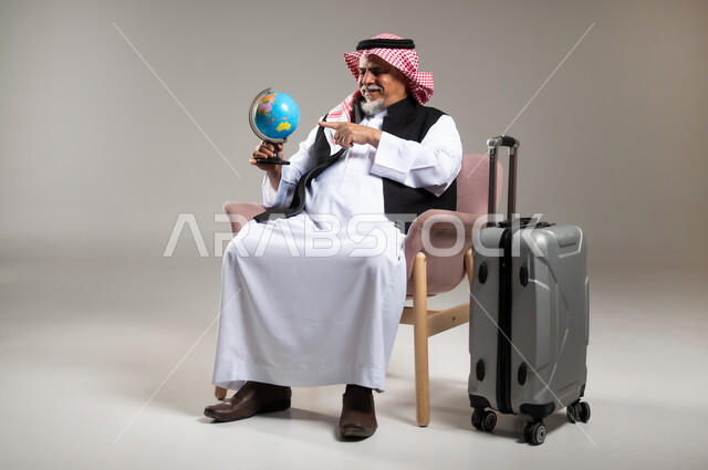 مفهوم البحث والاستكشاف حول العالم, الاشارة إلى مكان معين على الخريطة والنظر إلى أحد القارات, بورتريه لرجل عربي خليجي سعودي مبتسم كبير بالسن يرتدي الثوب التقليدي والشماغ يحمل بيده مجسم الكرة الارضية, الجلوس بجانب حقيبة الامتعة, خلفية رمادية