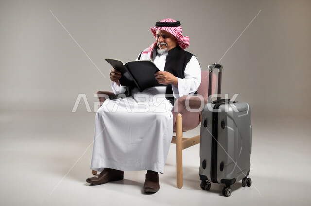 Preparing to have a good time on a tourist trip, practicing the hobby of reading and studying while waiting for the plane, portrait of an elderly Saudi Arabian Gulf man wearing the traditional thobe and shemagh sitting on a chair and holding a book in his hand next to a travel bag, reading carefully and with concentration, gray background