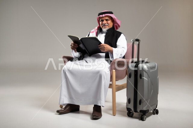 الاستعداد لقضاء وقت ممتع في الرحلات السياحية, ممارسة هواية القراءة والمطالعة في وقت انتظار الطائرة, بورتريه  لرجل كبير بالسن عربي سعودي خليجي يرتدي الثوب والشماغ التقليدي يجلس على الكرسي ويمسك بيده الكتاب بجانب حقيبة السفر, القراءة بتمعن وتركيز, خلفية رمادية