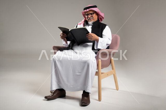 مفهوم محو الأمية والتعلم الذاتي, ممارسة هواية القراءة والمطالعة, بورتريه  لرجل كبير بالسن عربي سعودي خليجي يرتدي الثوب والشماغ التقليدي يجلس على الكرسي ويمسك بيده الكتاب, القراءة بتمعن وتركيز, خلفية رمادية