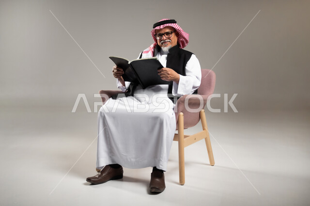 Reading and studying hobby, concept of literacy and self-learning, portrait of an elderly Saudi Arabian Gulf man wearing traditional thobe and shemagh sitting on a chair holding a book in his hand, reading carefully and with concentration, gray background