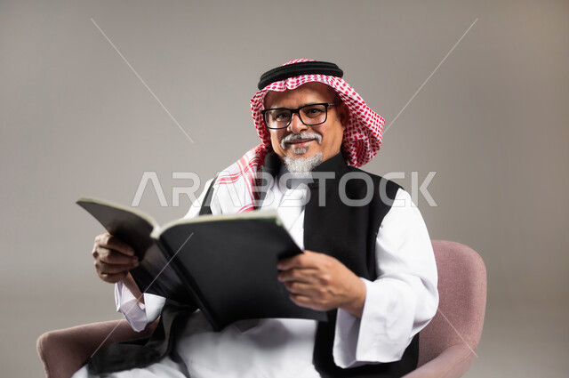 Concept of literacy and self-learning, practicing the hobby of reading and studying, close-up portrait of an elderly Saudi Arabian Gulf man wearing the traditional thobe and shemagh sitting on a chair and holding a book in his hand, reading carefully and with concentration, gray background