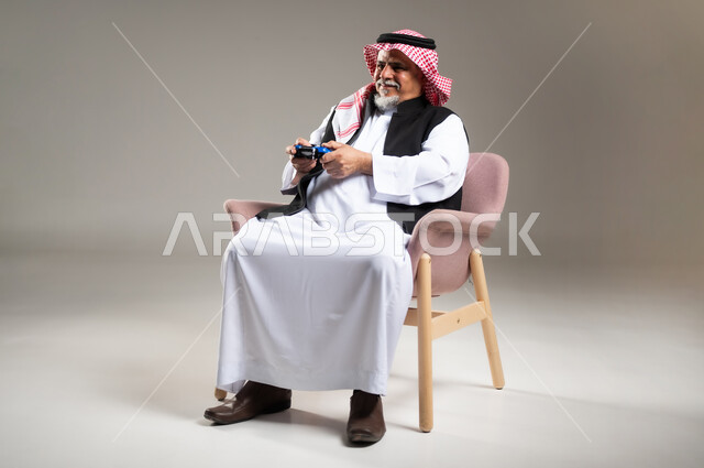 استخدام الأجهزة التكنولوجية والتقنيات الحديثة، بورتريه لرجل كبير بالسن عربي خليجي سعودي مبتسم يرتدي الثوب والشماغ التقليدي يجلس على الكرسي يمسك بيده عصا التحكم بإيماءات السرور والاندماج، تعابير الاستمتاع  باللعب بالألعاب الالكترونية الترفيهية، خلفية رمادية