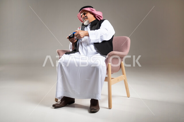 استخدام الأجهزة التكنولوجية والتقنيات الحديثة، بورتريه لرجل كبير بالسن عربي خليجي سعودي مبتسم يرتدي الثوب والشماغ التقليدي يجلس على الكرسي يمسك بيده عصا التحكم بإيماءات السرور والاندماج، تعابير الاستمتاع  باللعب بالألعاب الالكترونية الترفيهية، خلفية رمادية