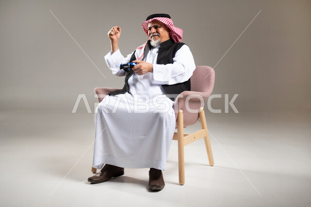 Expressions of enjoyment playing entertaining electronic games, using technological devices and modern techniques, portrait of an elderly smiling Saudi Arabian Gulf Arab man wearing the traditional thobe and shemagh sitting on a chair holding a joystick with gestures of encouragement and enthusiasm, gray background
