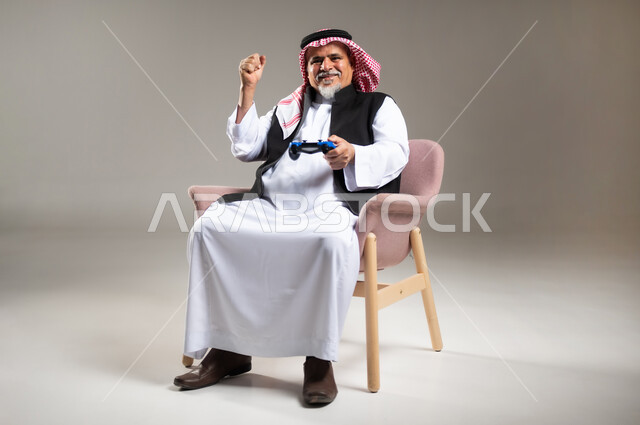 Enjoying playing entertaining electronic games, using technological devices and modern techniques, portrait of an elderly smiling Saudi Arabian Gulf Arab man wearing the traditional thobe and shemagh sitting on a chair holding a joystick with gestures of encouragement and enthusiasm, gray background