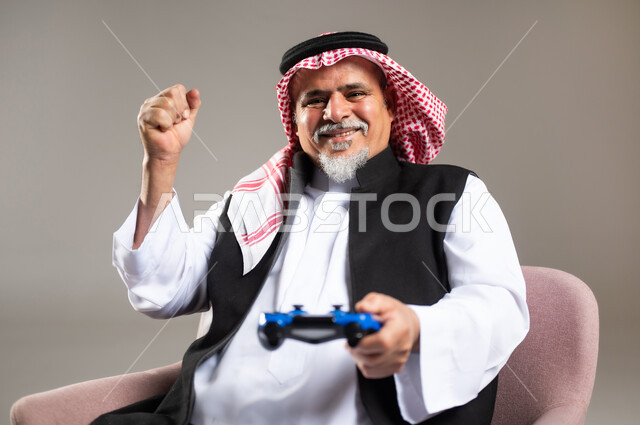 Enjoying playing entertaining electronic games, using technological devices and modern techniques, close-up portrait of an elderly smiling Saudi Arabian Gulf Arab man wearing the traditional thobe and shemagh sitting on a chair holding a joystick with gestures of encouragement and enthusiasm, gray background