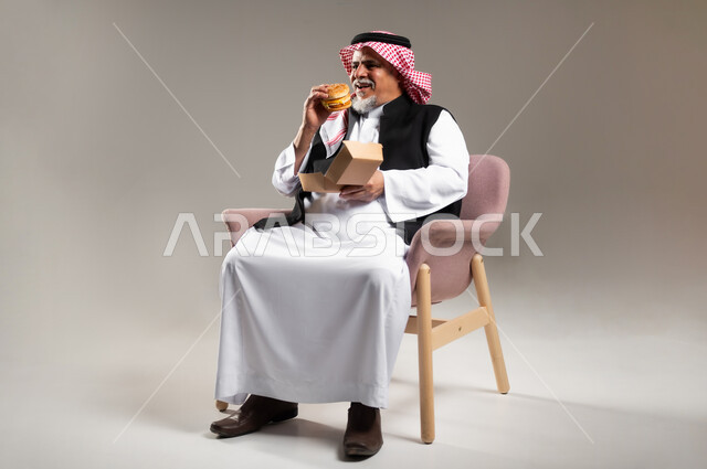 الاستمتاع بتناول الطعام الجاهز , مساوئ الإكثار من أكل الوجبات السريعة للكبار بالعمر , بورتريه لرجل عربي خليجي سعودي كبير في السن يرتدي الثوب التقليدي و الشماغ يجلس على الكرسي يقوم بأكل البرغر , صورة للجسم بالطول الكامل , خلفية رمادية
