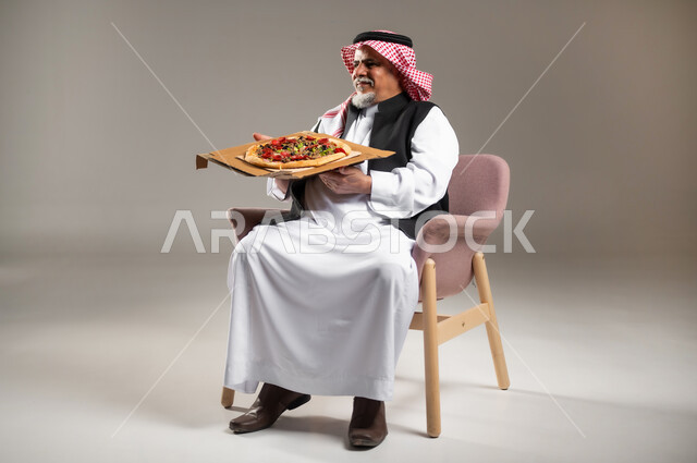 ضرر الإكثار من أكل الوجبات السريعة للكبار بالعمر , تناول طعام الغداء , بورتريه لرجل عربي خليجي سعودي كبير في السن يرتدي الثوب التقليدي و الشماغ يجلس على الكرسي يمسك بيده علبة البيتزا , صورة للجسم بالطول الكامل , خلفية رمادية