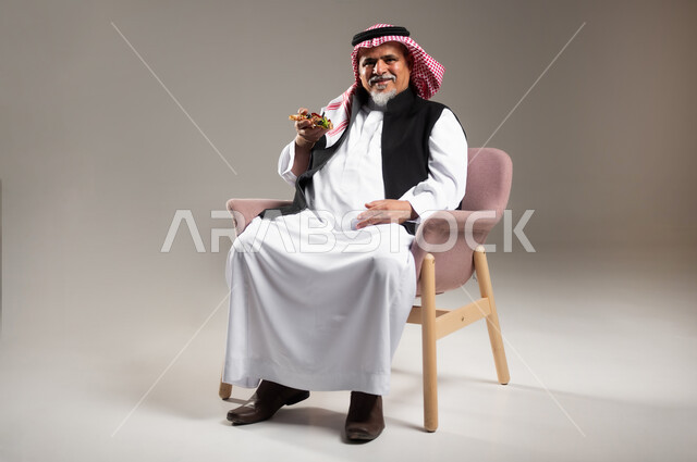 الاستمتاع بتناول الوجبات السريعة على الغداء , النظر غلى الكاميرا بإيماءات الفرح و السرور , بورتريه لرجل عربي خليجي سعودي كبير في السن يرتدي الثوب التقليدي و الشماغ يجلس على الكرسي و يمسك قطعة بيتزا بيده , صورة للجسم بالطول الكامل , خلفية رمادية