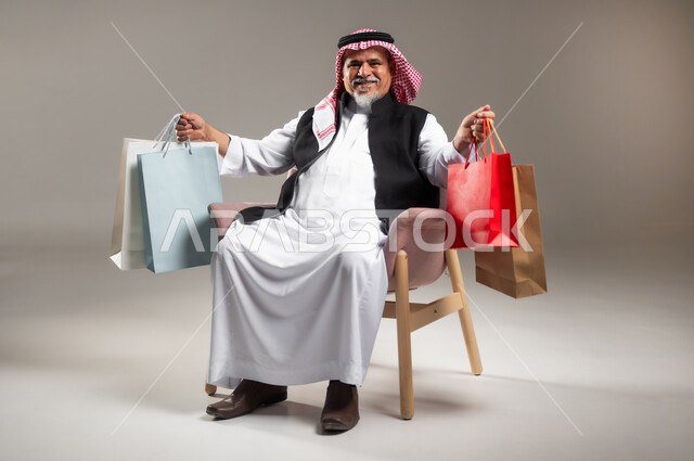مفهوم التسوق عن بعد , عمليات الشراء الالكتروني عبر الانترنت , بورتريه لرجل عربي خليجي سعودي كبير في السن يرتدي الثوب التقليدي و الشماغ يجلس على الكرسي يحمل أكياس مشتريات ورقية بيديه , صورة للجسم بالطول الكامل , خلفية رمادية