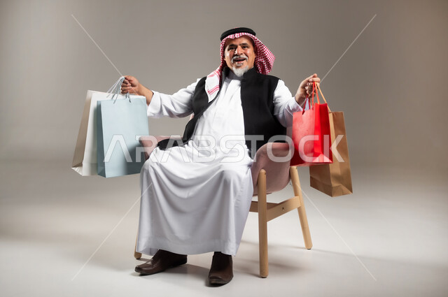عمليات الشراء الالكتروني عبر الانترنت , مفهوم التسوق عن بعد , بورتريه لرجل عربي خليجي سعودي كبير في السن يرتدي الثوب التقليدي و الشماغ يجلس على الكرسي يحمل أكياس مشتريات ورقية بيديه , صورة للجسم بالطول الكامل , خلفية رمادية