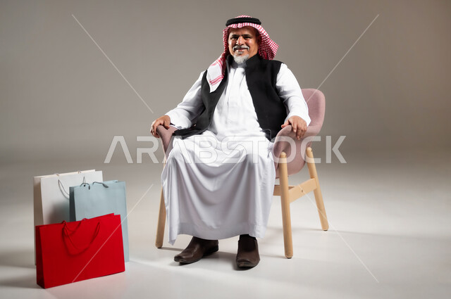 Providing means to help elderly people in the purchase and payment process remotely, the concept of electronic shopping, a portrait of an elderly Saudi Arabian Gulf man wearing a traditional thobe and shemagh sitting on a chair with paper bags next to him, a full-length portrait of the body, gray background.