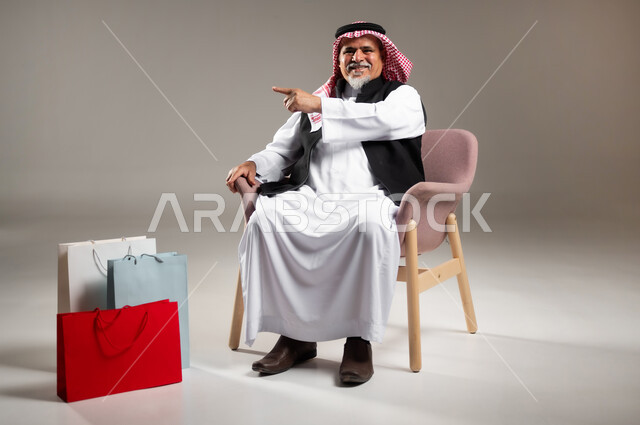 مفهوم الشراء عبر الانترنت , التسوق الالكتروني في السعودية , بورتريه لرجل عربي خليجي سعودي كبير في السن يرتدي الثوب التقليدي و الشماغ يجلس على الكرسي يشير بإصبع السبابة و بجانبه أكياس ورقية , صورة للجسم بالطول الكامل , خلفية رمادية