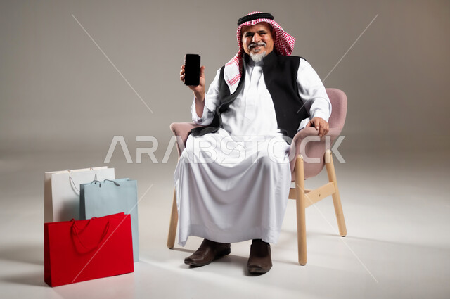 شراء المنتجات و الأغراض عبر الانترنت , بورتريه لرجل عربي خليجي سعودي كبير في السن يرتدي الثوب التقليدي و الشماغ يجلس على الكرسي يمسك الهاتف المحمول بيده و بجانبه أكياس ورقية , عرض شاشة سوداء فارغة عبر الجوال  , صورة للجسم بالطول الكامل , خلفية رمادية