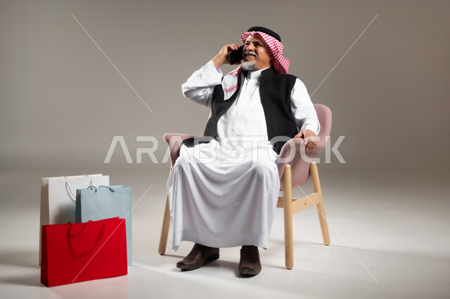 The concept of electronic shopping, making a phone call via mobile phone, portrait of an old Saudi Gulf Arab man wearing a traditional dress and shemagh, sitting on a chair holding a mobile phone in his hand and paper bags next to him, full-length body portrait, gray background.