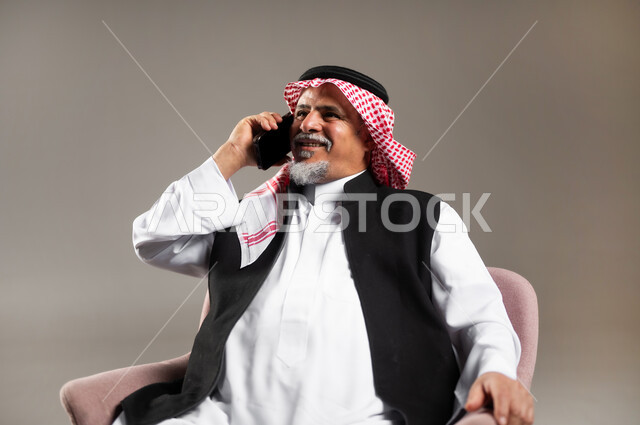 إجراء اتصال هاتفي عبر الجوال , التواصل مع العائلة و الأصدقاء في أوقات الفراغ , مشاركة الأخبار الجيدة , بورتريه مقرب لرجل عربي خليجي سعودي كبير في السن يرتدي الثوب التقليدي و الشماغ يجلس على الكرسي يمسك الهاتف المحمول بيده , خلفية رمادية