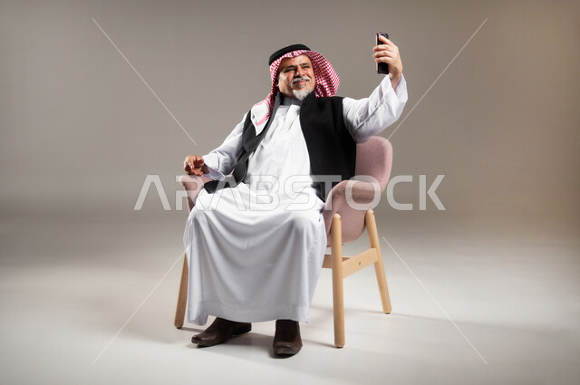 Making a video call via mobile phone, communicating with family and friends in free time, portrait of an elderly Saudi Gulf Arab man wearing a traditional thobe and shemagh, sitting on a chair and holding a mobile phone in his hand, full-length body portrait, gray background.