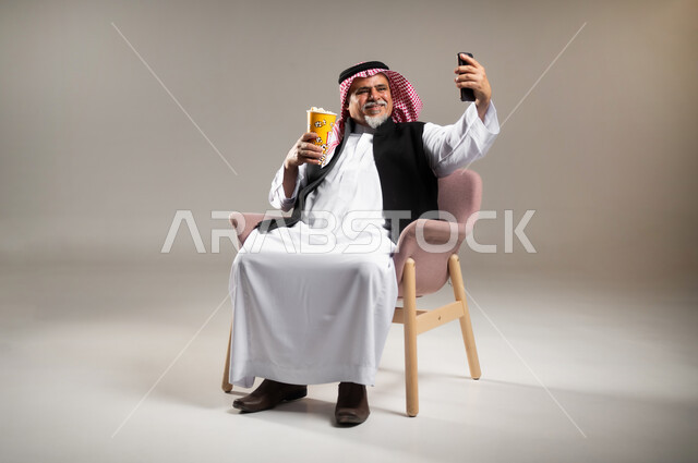 التقاط الصور التذكارية بواسطة الجوال , استخدام التكنولوجيا المتطورة للتسلية و الترفيه , بورتريه لرجل عربي خليجي سعودي كبير في السن يرتدي الثوب التقليدي و الشماغ يجلس على الكرسي يمسك بيديه الهاتف الممول و الفشار , صورة للجسم بالطول الكامل , خلفية رمادية