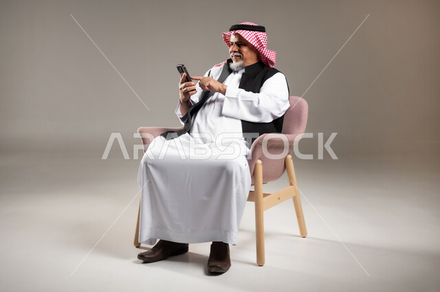 Communicating with friends via mobile device, using advanced devices for fun and entertainment, portrait of an elderly Saudi Gulf Arab man wearing a traditional thobe and shemagh, sitting on a chair and holding a mobile phone in his hand, full-length body portrait, gray background.