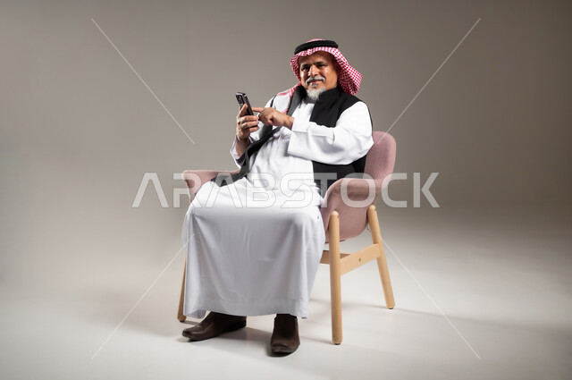 Browsing social media, using modern technical devices for fun and entertainment, portrait of an old Saudi Gulf Arab man wearing a traditional thobe and shemagh, sitting on a chair holding a mobile phone in his hand, full-length body portrait, gray background.