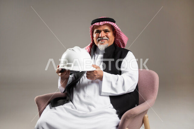 النظر إلى الكاميرا بإيماءات الفرح و السرور , العمل في مجال الهندسة المعمارية , مهن و وظائف في السعودية , بورتريه مقرب لرجل عربي خليجي سعودي كبير في السن يرتدي الثوب التقليدي و الشماغ يجلس على الكرسي و يمسك خوذة الرأس بيديه , خلفية رمادية