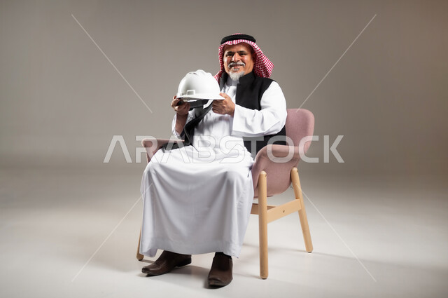 Work in the field of architecture, professions and jobs in Saudi Arabia, portrait of an old Saudi Gulf Arab man wearing the traditional dress and shemagh, sitting on a chair holding a helmet in his hand, looking at the camera with gestures of joy and pleasure, full-length body photo, gray background.