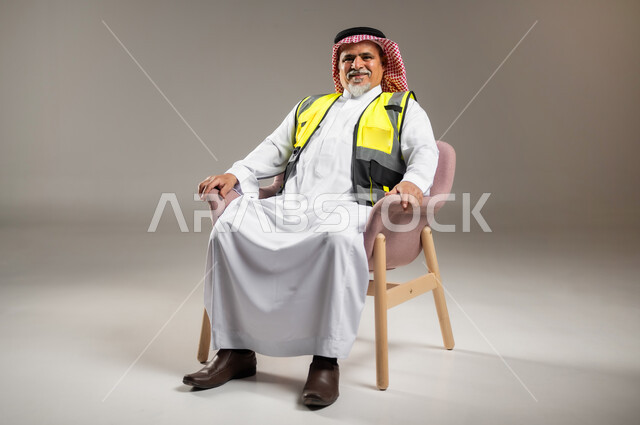 النظر إلى الكاميرا بإيماءات السعادة و السرور , حب العمل في مجال الهندسة المعمارية , بورتريه لمهندس عربي خليجي سعودي كبير في السن يرتدي الثوب التقليدي و الشماغ مع سترة الحماية يجلس على الكرسي , صورة للجسم بالطول الكامل , خلفية رمادية