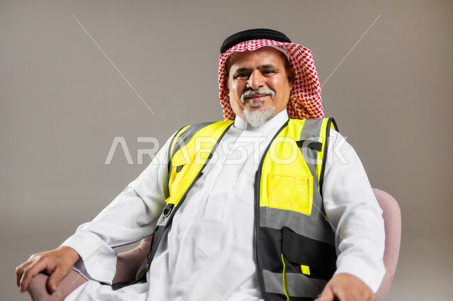 حب العمل في مجال الهندسة المعمارية , مهن و وظائف هندسية في السعودية  , بورتريه مقرب لمهندس عربي خليجي سعودي كبير في السن يرتدي الثوب التقليدي و الشماغ مع سترة الحماية يجلس على الكرسي , النظر إلى الكاميرا بإيماءات الفرح و السرور , خلفية رمادية