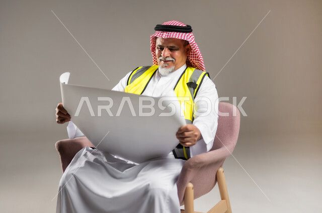 إدارة الأعمال و المشاريع الهندسية , مفهوم الهندسة و الإنشاءات المعمارية , بورتريه مقرب لمهندس عربي خليجي سعودي كبير في السن يرتدي الثوب التقليدي و الشماغ مع سترة الحماية يجلس على الكرسي و يمسك بيده المخططات , خلفية رمادية