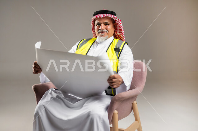 مراجعة الأعمال و المخططات الهندسية , العمل في المجال الهندسي , بورتريه مقرب لمهندس عربي خليجي سعودي كبير في السن يرتدي الثوب التقليدي و الشماغ مع سترة الحماية يجلس على الكرسي و يمسك بيده  المخططات , خلفية رمادية