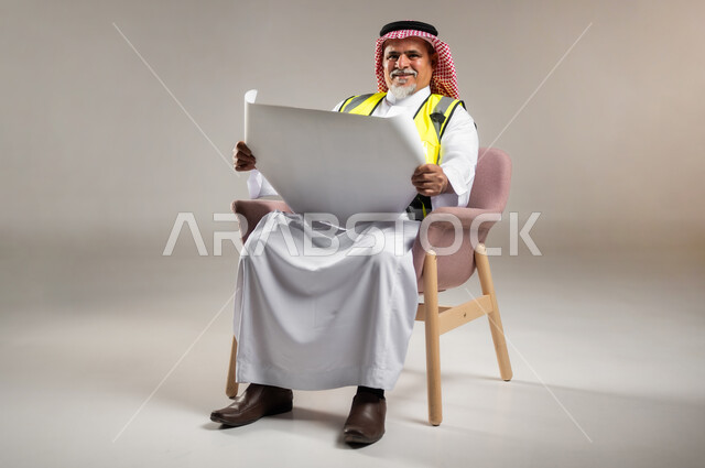 الإشراف على الأعمال الهندسية و مخططات البناء , مهن و وظائف في السعودية , بورتريه لمهندس عربي خليجي سعودي كبير في السن يرتدي الثوب التقليدي و الشماغ مع سترة الحماية يجلس على الكرسي و يمسك بيده المخططات , صورة للجسم بالطول الكامل , خلفية رمادية