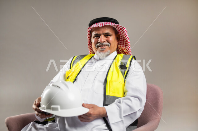 مفهوم مهنة الهندسة المعمارية , الإعمار بسواعد أبناء الوطن , بورتريه مقرب لمهندس عربي خليجي سعودي كبير في السن يرتدي الثوب التقليدي و الشماغ مع سترة الحماية يجلس على الكرسي و يمسك بيده خوذة الرأس , النظر إلى الكاميرا بإيماءات السعادة و السرور , خلفية رمادية