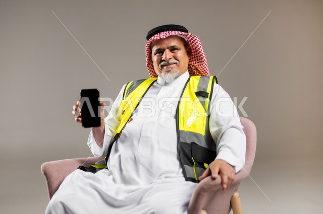 Using advanced devices at work, the concept of engineering and architectural construction, a close-up portrait of an elderly Saudi Arabian Gulf engineer wearing a traditional thobe and shemagh with a protective jacket sitting on a chair and holding a mobile phone in his hand, showing a blank black screen on the mobile phone, gray background