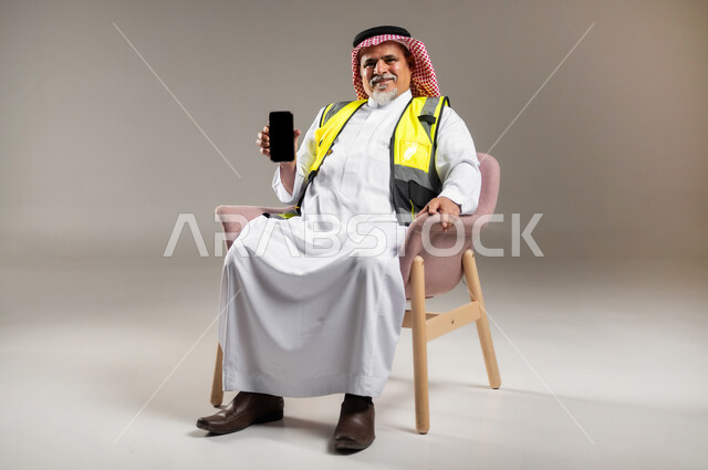 استخدام الأجهزة التقنية الحديثة , العمل في القطاع الهندسي , بورتريه لمهندس عربي خليجي سعودي كبير في السن يرتدي الثوب التقليدي و الشماغ مع سترة الحماية يجلس على الكرسي و يمسك بيده الهاتف المحمول , عرض شاشة سوداء فارغة عبر الجوال , صورة للجسم بالطول الكامل , خلفية رمادية