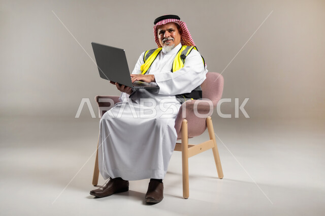 Integrating modern technology with engineering work, using a computer to accomplish tasks, architectural concept, portrait of an elderly Saudi Arabian Gulf engineer wearing a traditional thobe and shemagh with a protective jacket sitting on a chair and holding a laptop in his hand, full-length body portrait, gray background.