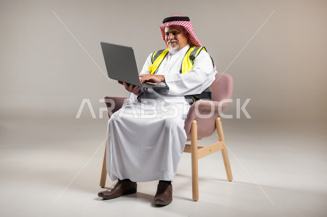 Using a computer to accomplish tasks, integrating modern technologies into engineering work, engineering concept for an architecture, portrait of an elderly Saudi Arabian Gulf engineer wearing a traditional thobe and shemagh with a protective jacket sitting on a chair and holding a laptop in his hand, a full-length portrait of the body, gray background.