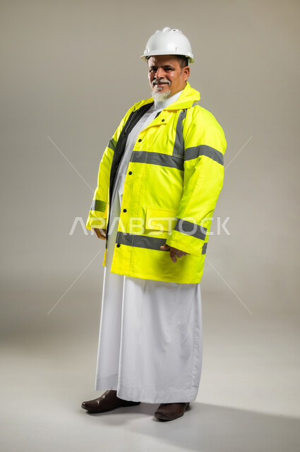 الإعمار بسواعد أبناء الوطن , العمل في القطاع الهندسي , بورتريه لمهندس عربي خليجي سعودي كبير في السن يرتدي الثوب التقليدي مع السترة و خوذة الحماية , النظر إلى الكاميرا بإيماءات السعادة و السرور , صورة للجسم بالطول الكامل , خلفية رمادية