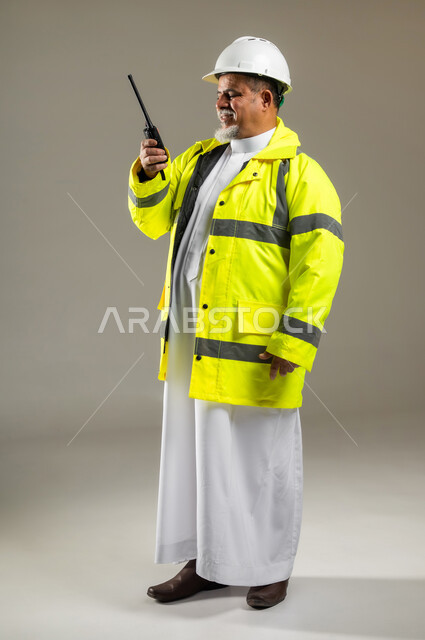 مفهوم الهندسة المعمارية , استخدام جهاز الاتصال اللاسلكي في العمل , بورتريه لمهندس عربي خليجي سعودي كبير في السن يرتدي الثوب التقليدي مع السترة و خوذة الحماية , صورة للجسم بالطول الكامل , خلفية رمادية