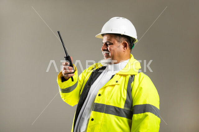 مهن و وظائف هندسية في السعودية , استخدام جهاز الاتصال اللاسلكي في العمل , مفهوم الهندسة و الإنشاءات المعمارية , بورتريه مقرب لمهندس عربي خليجي سعودي كبير في السن يرتدي الثوب التقليدي مع السترة و خوذة الحماية , خلفية رمادية