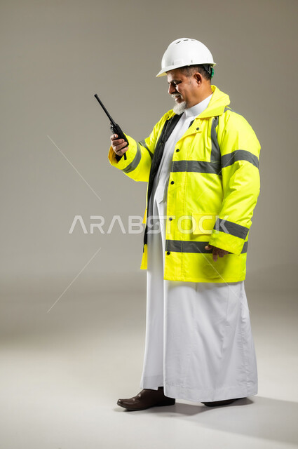 مفهوم الهندسة و الإنشاءات المعمارية , استخدام جهاز الاتصال اللاسلكي في العمل , بورتريه لمهندس عربي خليجي سعودي كبير في السن يرتدي الثوب التقليدي مع السترة و خوذة الحماية , صورة للجسم بالطول الكامل , خلفية رمادية