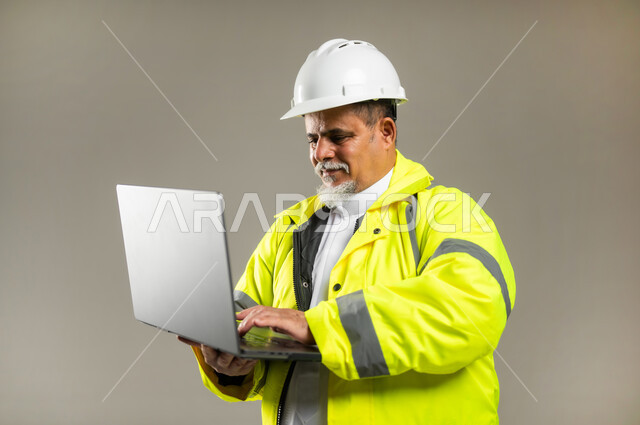 إنجاز المهام على جهاز الحاسوب , دمج التقنيات الحديثة بالأعمال الهندسية , العمل في القطاع الهندسي , بورتريه مقرب لمهندس عربي خليجي سعودي كبير في السن يرتدي الثوب التقليدي مع السترة و خوذة الحماية يحمل الحاسب المحمول بيديه , خلفية رمادية