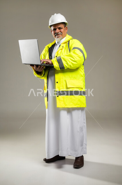 دمج التقنيات الحديثة بالأعمال الهندسية , مهن و وظائف في السعودية , بورتريه لمهندس عربي خليجي سعودي كبير في السن يرتدي الثوب التقليدي مع السترة و خوذة الحماية يحمل جهاز الحاسوب بيديه , صورة للجسم بالطول الكامل , خلفية رمادية
